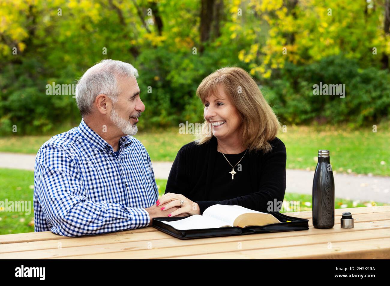 Una coppia matura che condivide insieme il tempo devozionale e studia la bibbia a un tavolo da picnic in una calda giornata di autunno in un parco cittadino Foto Stock