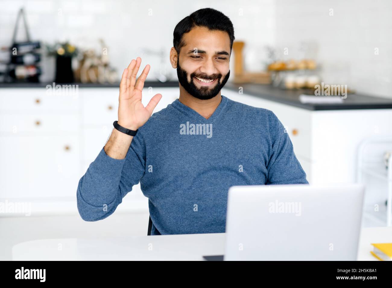 Il freelance indiano utilizza la connessione video per incontri virtuali con i colleghi, lavorando in remoto da casa, si siede di fronte al portatile in cucina, salutando e sventolando nella webcam Foto Stock