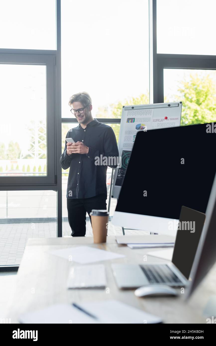 Programmatore positivo che utilizza smartphone vicino a lavagna a fogli mobili e computer in ufficio Foto Stock