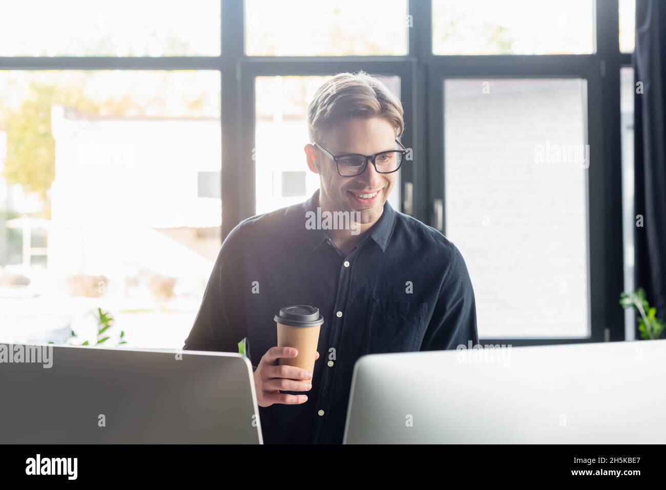 Programmatore sorridente con caffè per andare a guardare i monitor del computer in ufficio Foto Stock