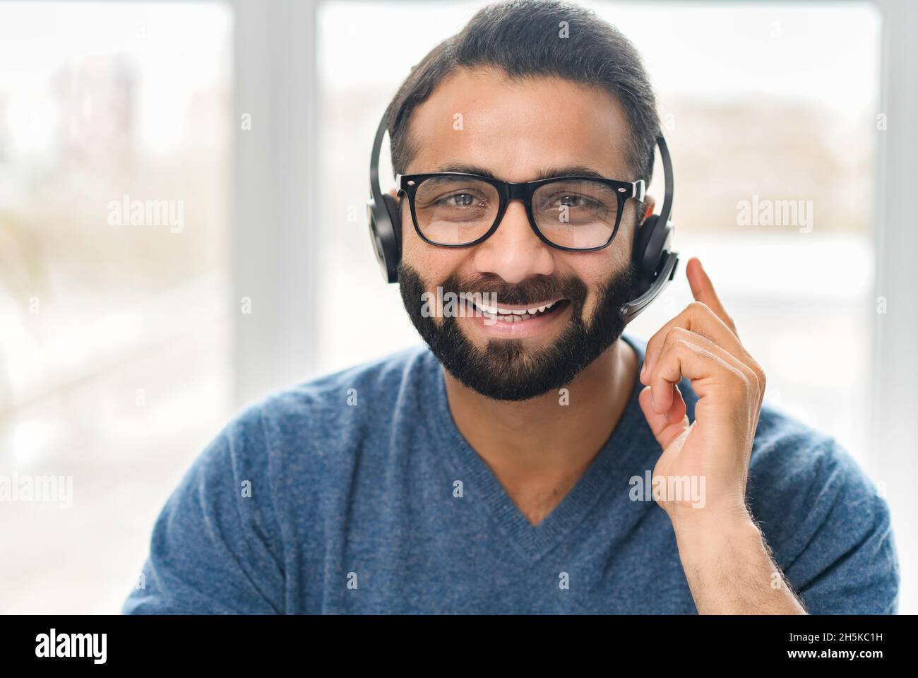 Addetto all'assistenza clienti che indossa un auricolare. Fidato uomo indiano dipendente in cuffie con microfono guarda la fotocamera con sorriso amichevole. Chat video con un operatore di call center orientale Foto Stock
