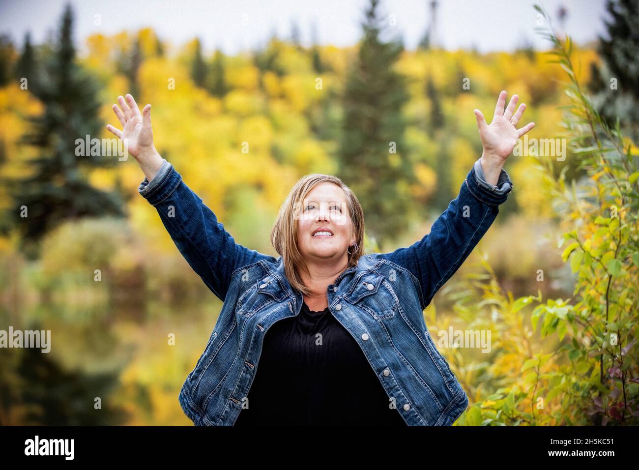 Una donna che gode di tempo di qualità all'aperto e che prende il tempo di alzare le mani in adorazione in un parco cittadino con un lago sullo sfondo Foto Stock
