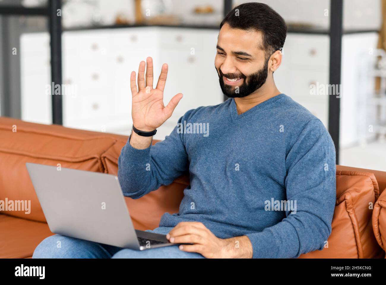 Cheerful dipendente indiano maschio saluto partecipanti di riunione virtuale, uomo multirazziale in elegante casual si siede di fronte al laptop e di wonding ciao in webcam, ha video conferenza con i colleghi Foto Stock