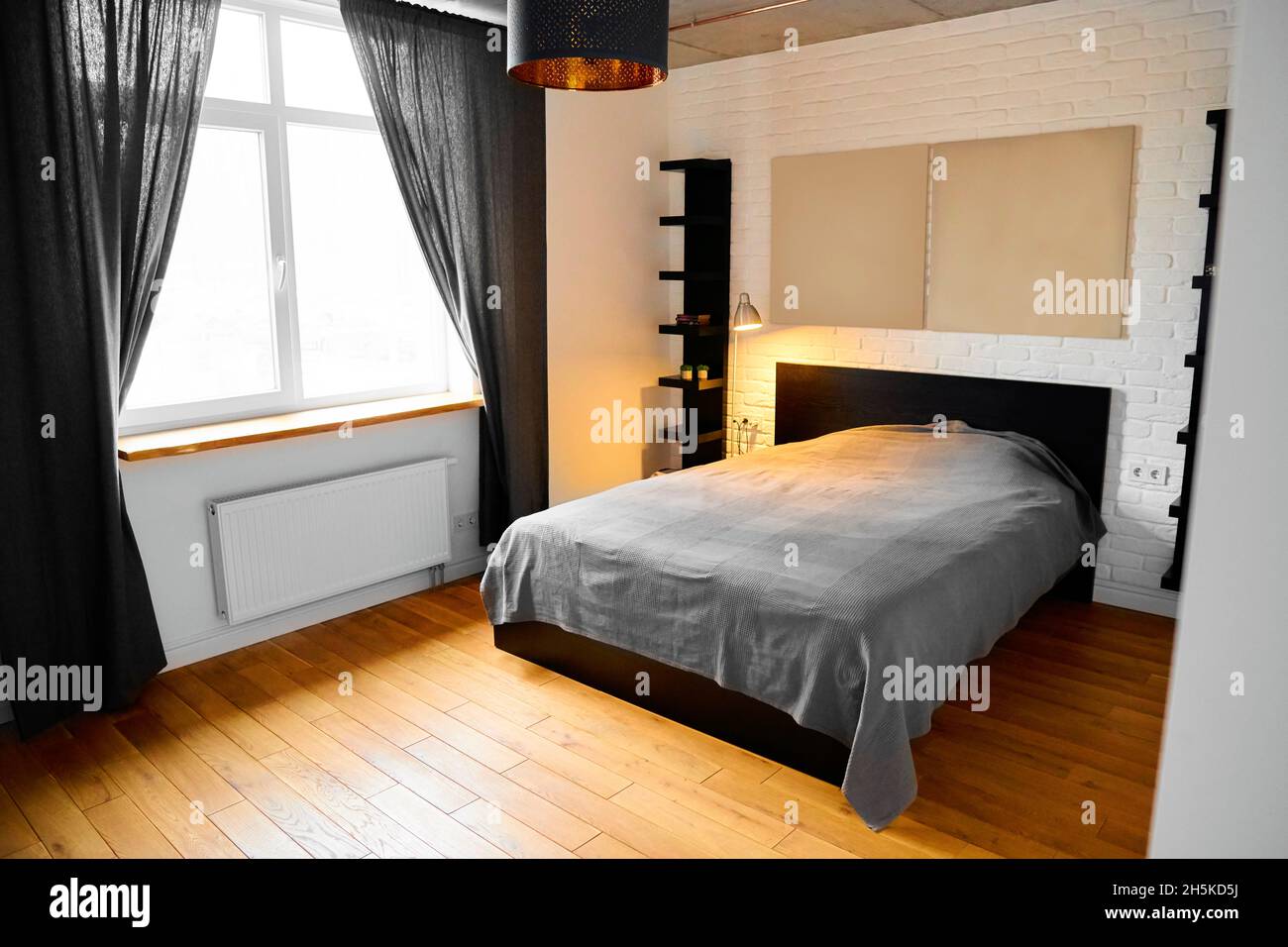 Camera da letto semplice in design industriale con pareti in mattoni bianchi, pavimento in legno, soffitto in cemento, tende in cotone nero e testiera letto a schermo intero in stile moderno Foto Stock