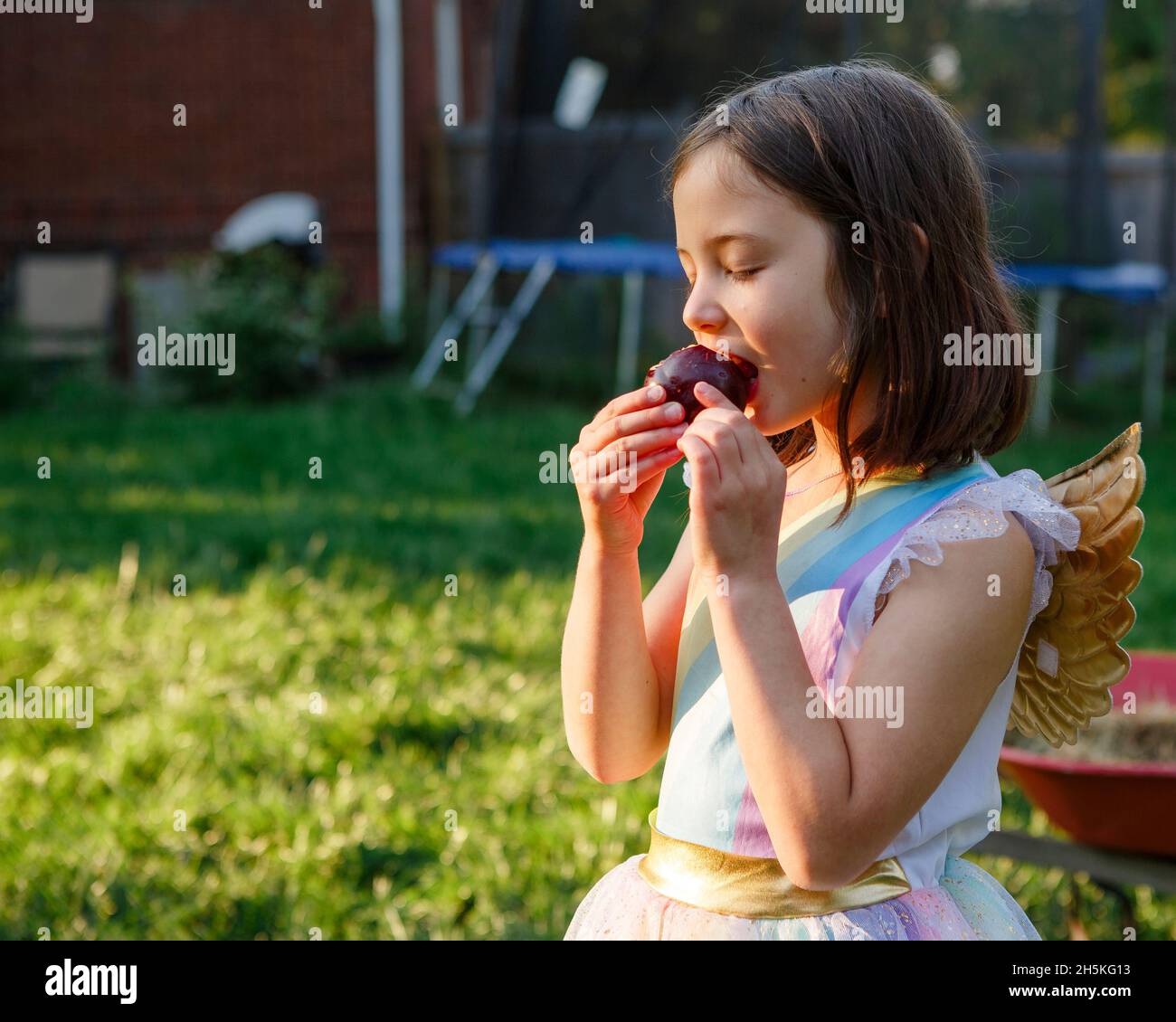 Un bambino piccolo in costume con ali dorate mangia succosa prugna sotto il sole Foto Stock