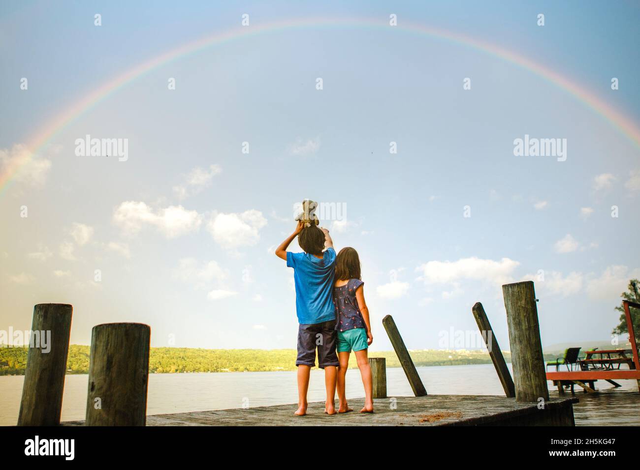 due bambini si levano in piedi sul ponte sotto l'arcobaleno vicino al lago illuminato dal sole con l'orso giocattolo Foto Stock