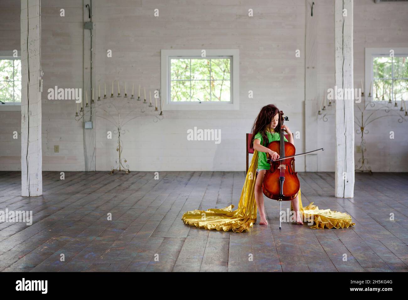 La ragazza Barefoot in vimini e mantello d'oro suona violoncello in un grande fienile vuoto Foto Stock