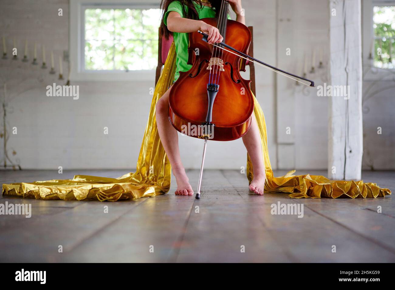 Metà inferiore di bambino a piedi nudi in oro mantello giocando violoncello in camera vuota Foto Stock