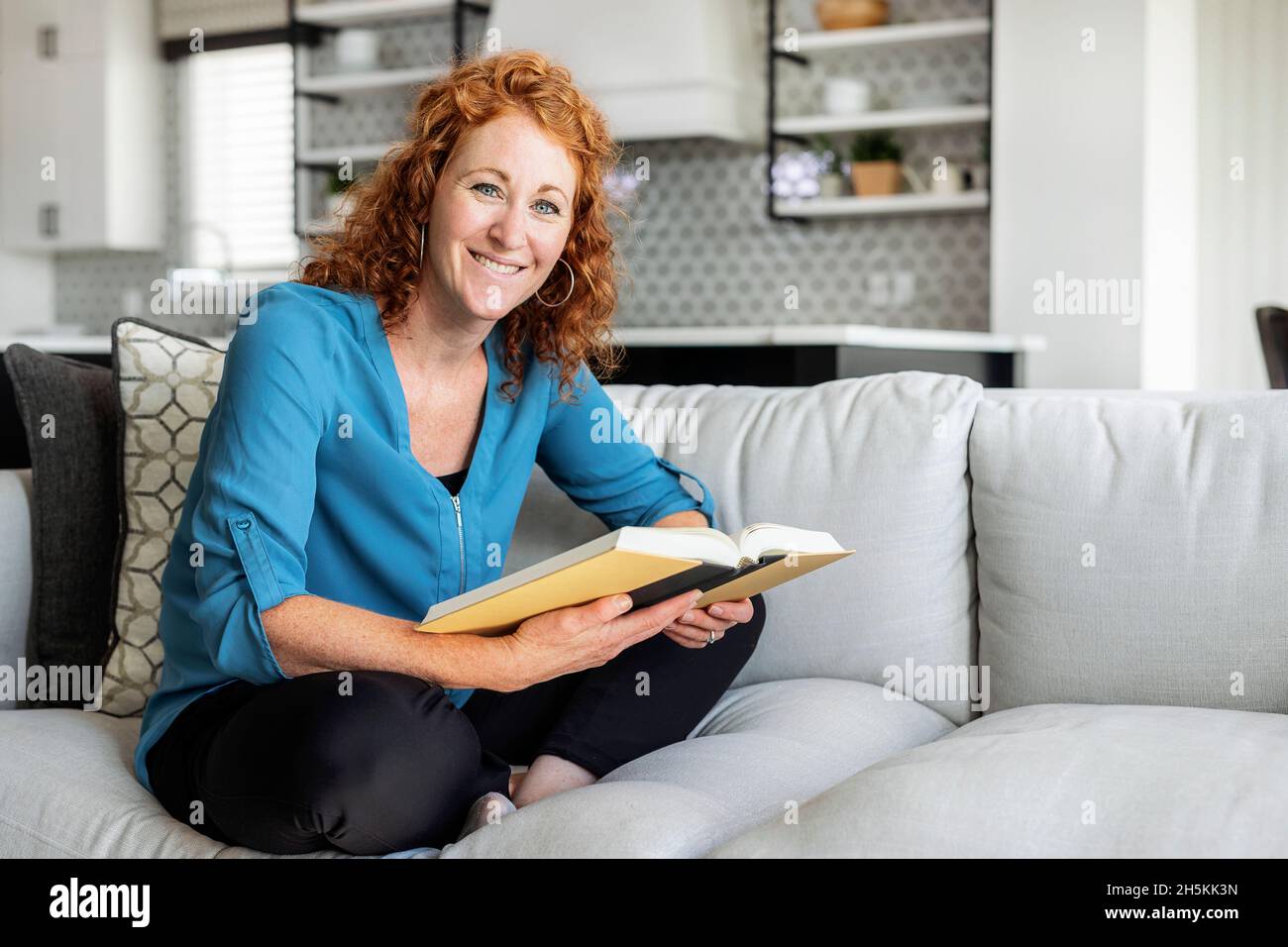 Una donna di metà adulta siede su un divano a casa leggendo un libro e guardando la macchina fotografica; Edmonton, Alberta, Canada Foto Stock