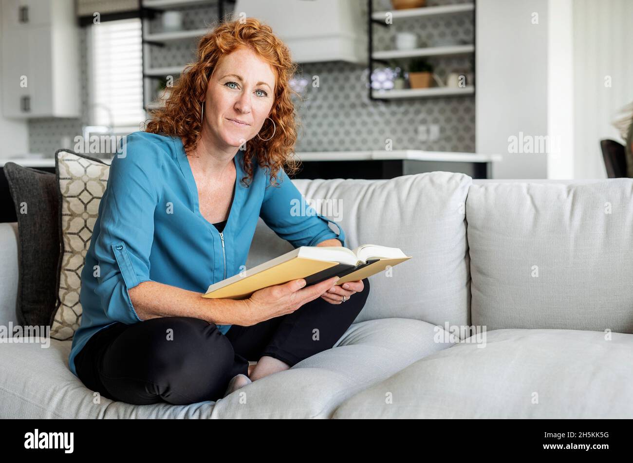 Una donna di metà adulta siede su un divano a casa leggendo un libro e guardando la macchina fotografica; Edmonton, Alberta, Canada Foto Stock