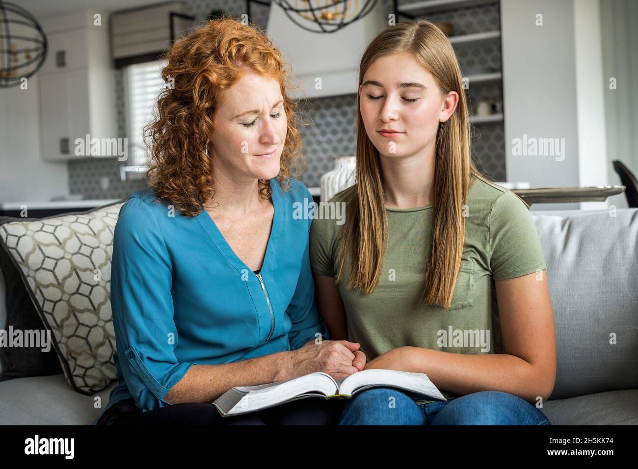 Madre e figlia adolescente seduti su un divano a casa leggendo la Bibbia e pregando insieme; Edmonton, Alberta, Canada Foto Stock