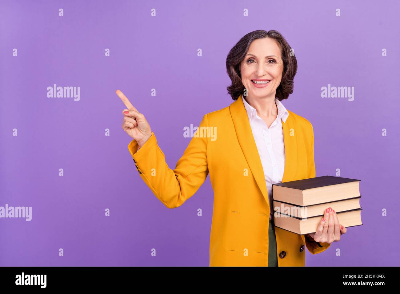 Foto di divertente brunette anziana signora indice spazio vuoto tenere libri indossare giacca gialla isolato su sfondo di colore viola Foto Stock