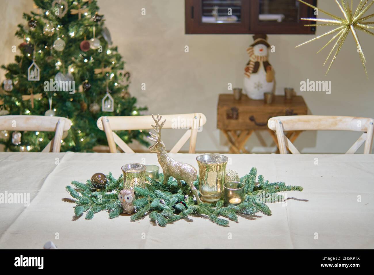 tavolo per cena di natale con bauble verde con renna dorata e candele. albero di natale decorato, stella dorata e statuetta di pupazzo di neve nel b Foto Stock