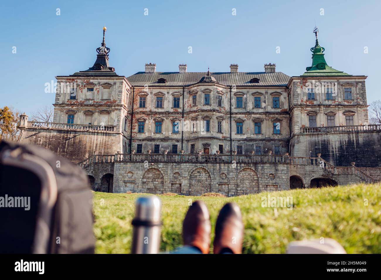 Il turista gode di vista sul castello di Pidhirtsi adagiato sull'erba. Viaggia verso luoghi storici e luoghi di interesse. Primo piano di scarpe e zaino con pallone sottovuoto. Foto Stock