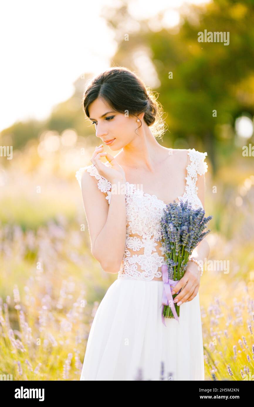 Sibenik, Croazia - 05.06.17: Sposa in un abito in pizzo bianco si erge in un campo e tiene in mano un bouquet di lavanda legato con un nastro di lilla, girando Foto Stock