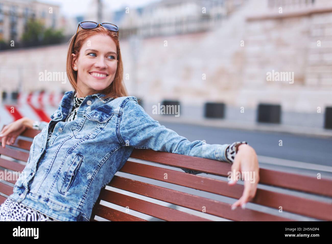 Felice giovane rossa caucasica positiva donna seduta su panchina in città con un sorriso toothy Foto Stock