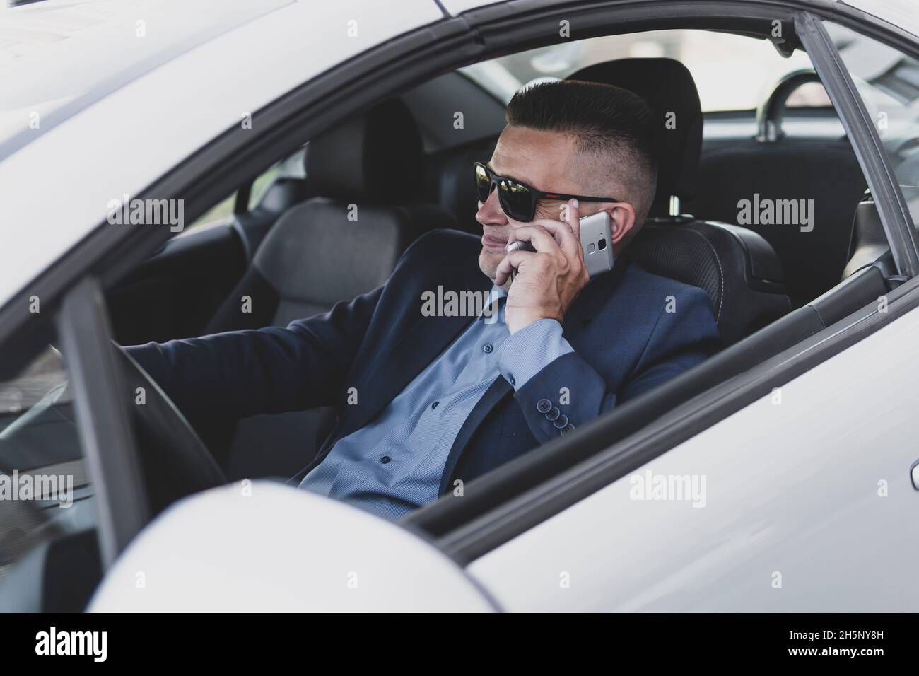 Ritratto di un bel uomo d'affari che parla al telefono seduto sul sedile dell'auto e sorridendo felicemente. Il conducente comunica su uno smartphone durante la guida. Busi Foto Stock