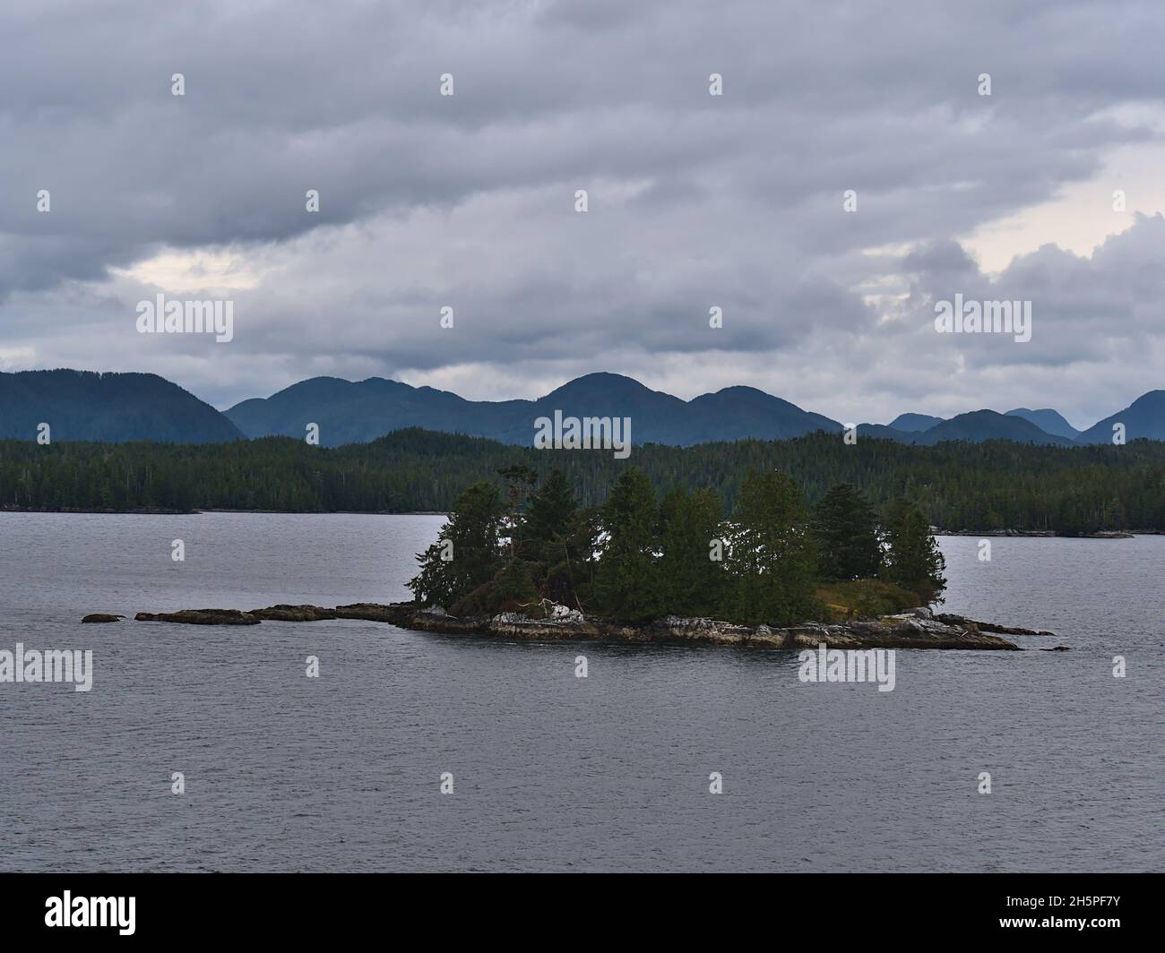 Piccola isola rocciosa coperta da vegetazione verde nel passaggio interno di fronte a Cunningham Island e Coast Mountains in British Columbia, Canada. Foto Stock