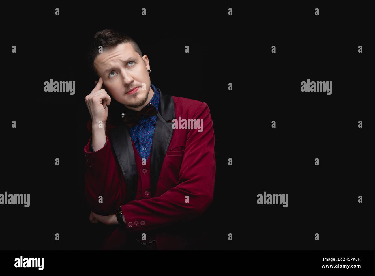 L'uomo con il piercing nelle orecchie in vestito rosso pone pensieroso, analizza la situazione. Sfondo nero isolato. Foto Stock