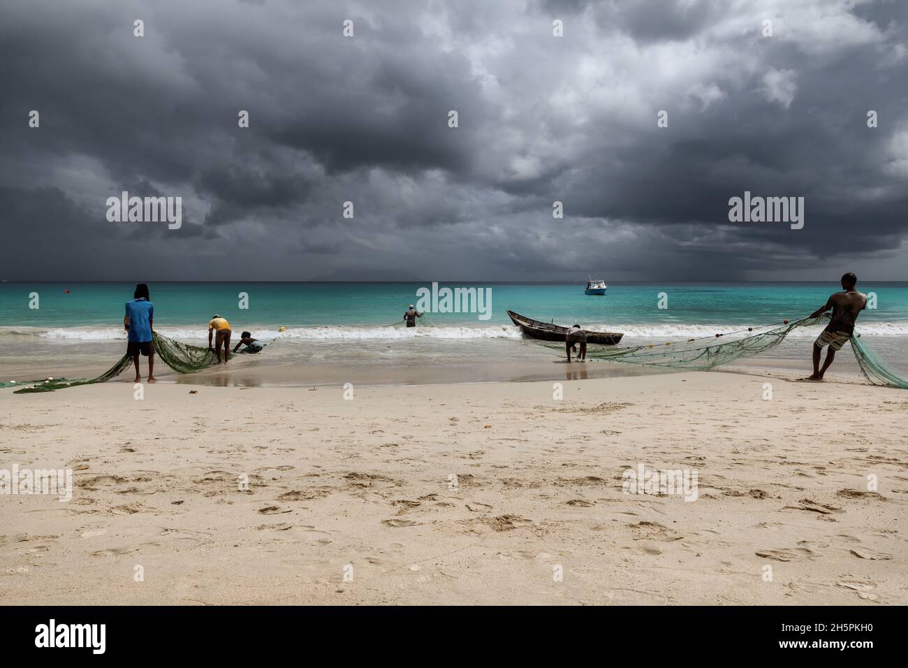 I pescatori tirano le reti dal mare su una spiaggia nei tropici con il cielo drammatico su Mahe Island, Seychelles Foto Stock