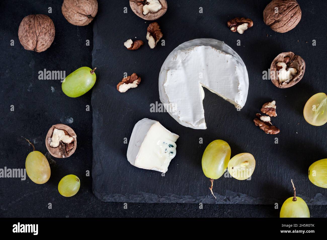 Tipo di formaggio Brie. Formaggio morbido con uva e noci su sfondo nero. Disposizione piatta, vista dall'alto Foto Stock
