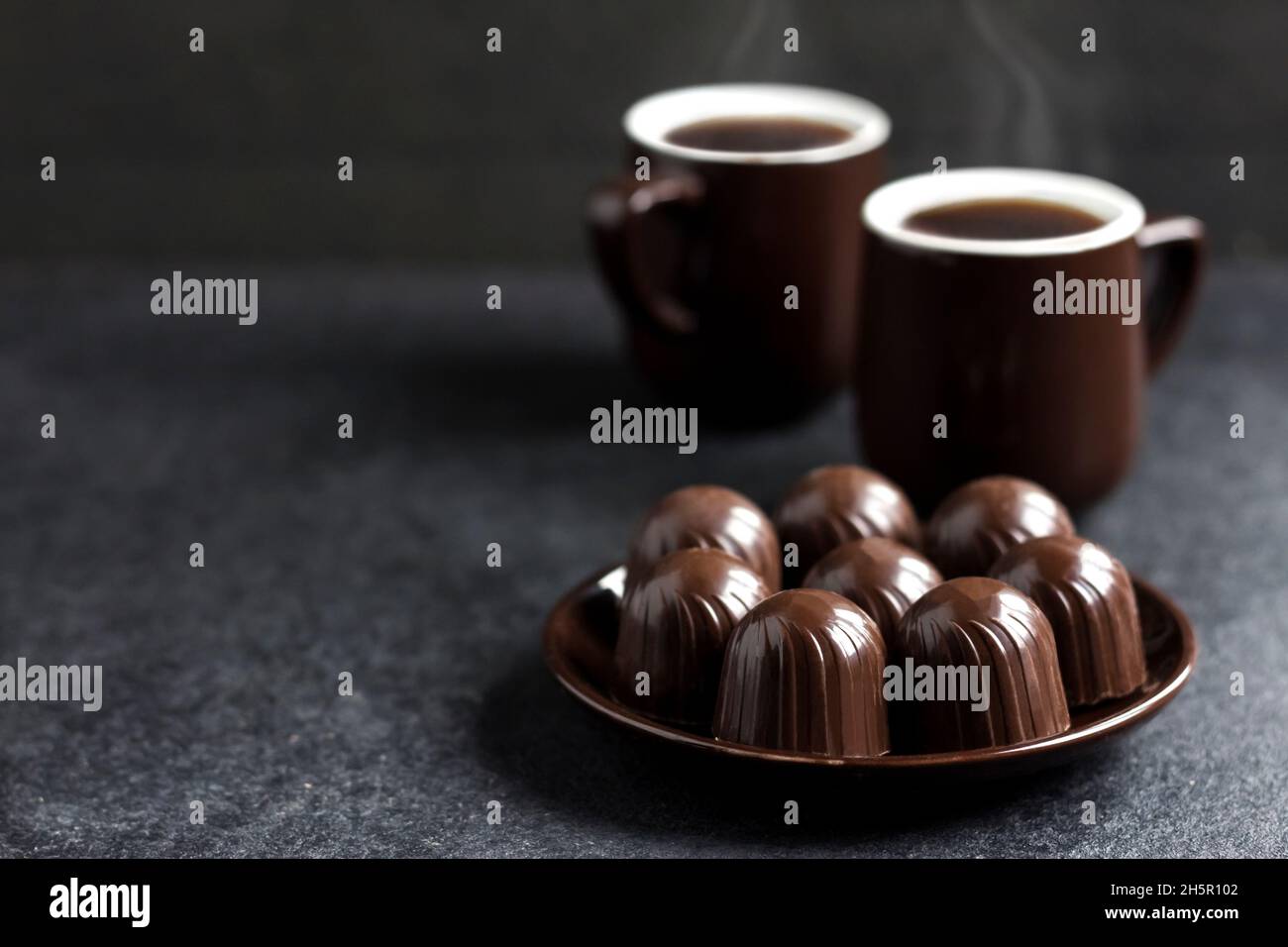 Caramelle al cioccolato su un piatto e due tazze di caffè caldo su sfondo nero con spazio copia Foto Stock