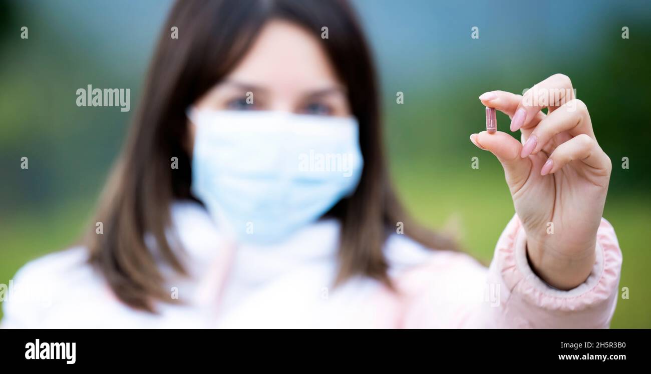 (Fuoco selettivo) Una giovane ragazza defocused che indossa una maschera chirurgica del viso sta tenendo una pillola rossa. Concetto di farmaco antivirale contro Covid-19. Foto Stock