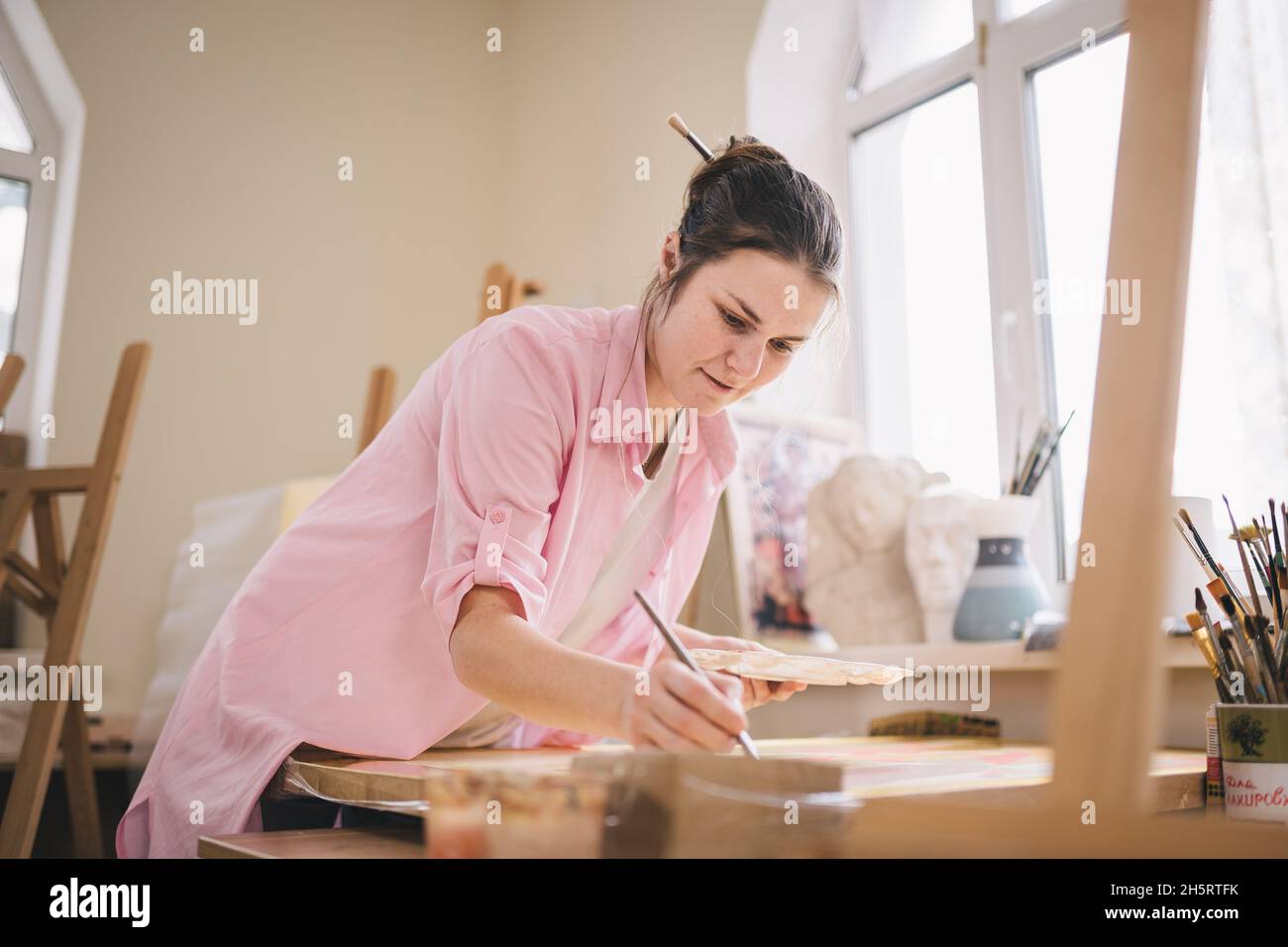 Artista donna caucasica che lavora a un dipinto in uno studio luminoso. Happy artista disegna un progetto d'arte con pitture e un pennello in officina Foto Stock