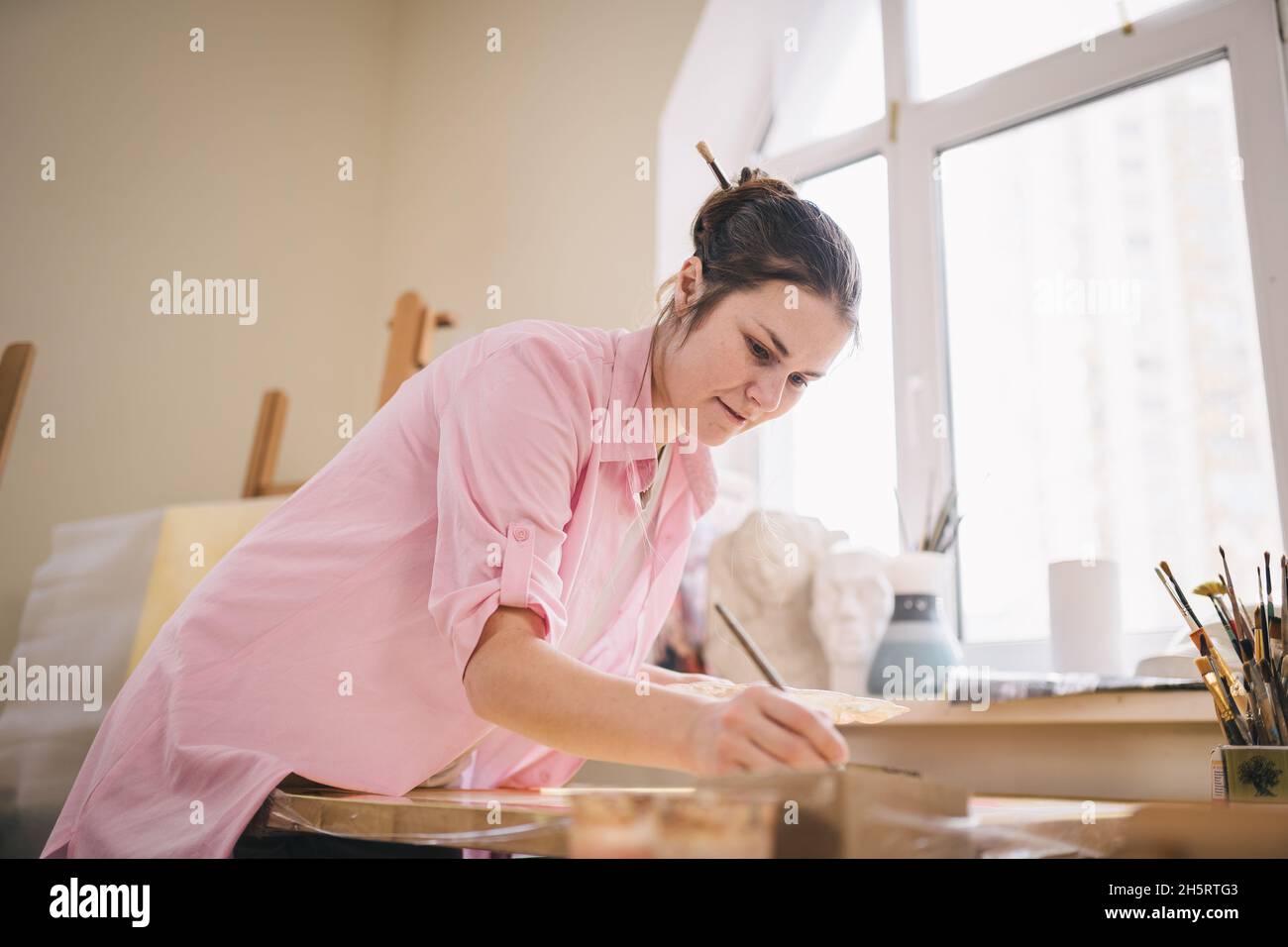 Cute donna dipinge su tela in un laboratorio d'arte. Artista che crea l'immagine. Scuola d'arte o studio. Lavorare con vernici, spazzole e cavalletto. Hobby e tempo libero Foto Stock