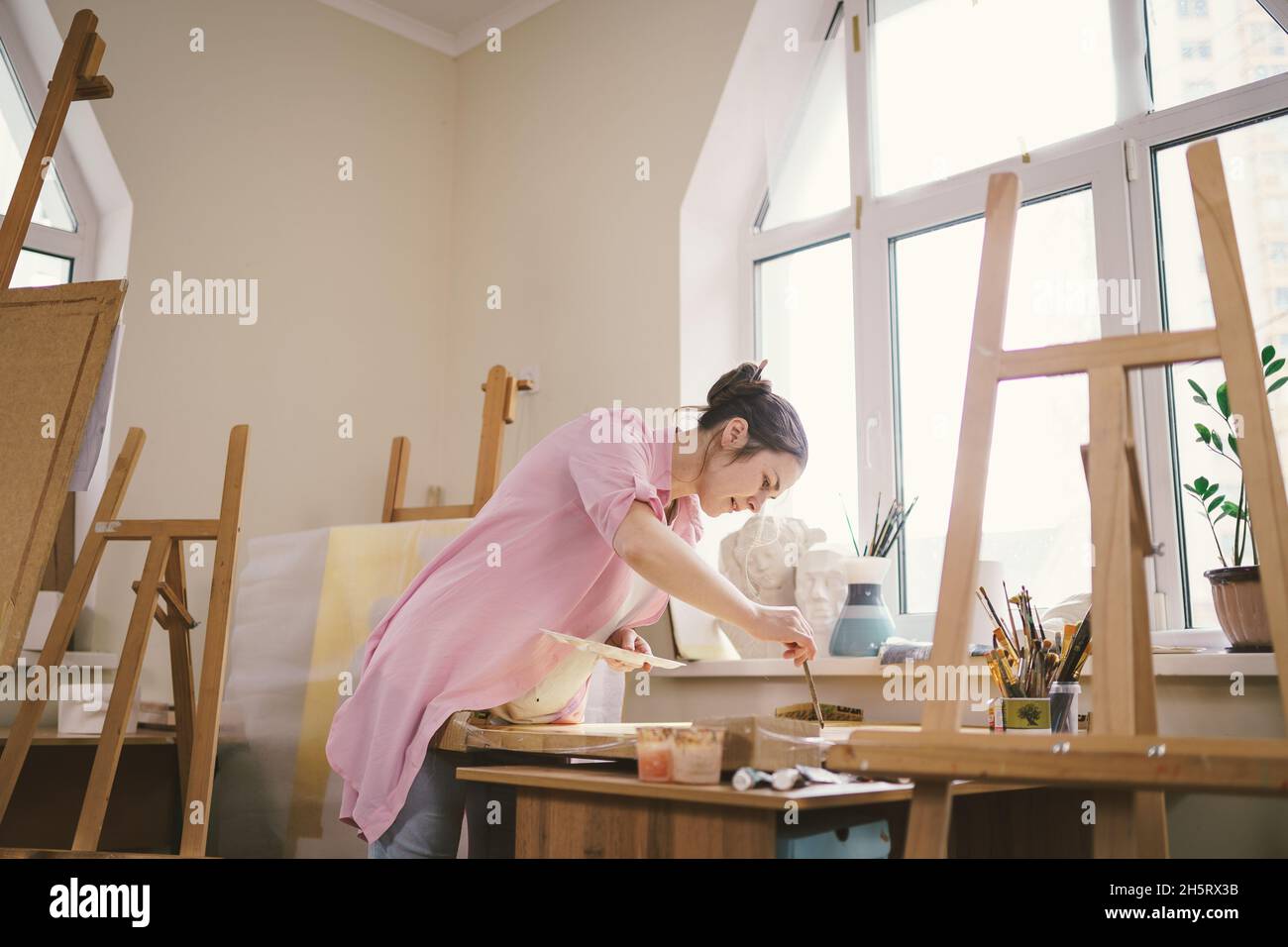 Artista donna caucasica che lavora a un dipinto in uno studio luminoso. Happy artista disegna un progetto d'arte con pitture e un pennello in officina Foto Stock