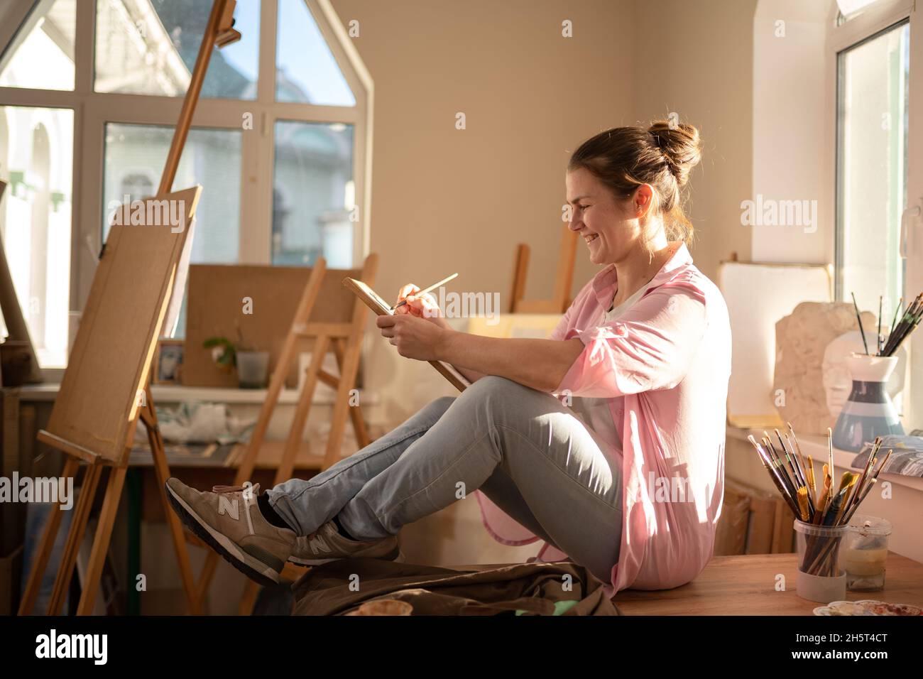 Cute donna dipinge su tela in un laboratorio d'arte. Artista che crea l'immagine. Scuola d'arte o studio. Lavorare con vernici, spazzole e cavalletto. Hobby e tempo libero Foto Stock