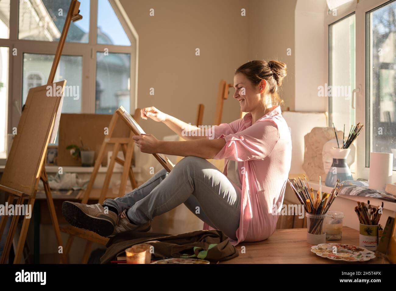 Cute donna dipinge su tela in un laboratorio d'arte. Artista che crea l'immagine. Scuola d'arte o studio. Lavorare con vernici, spazzole e cavalletto. Hobby e tempo libero Foto Stock