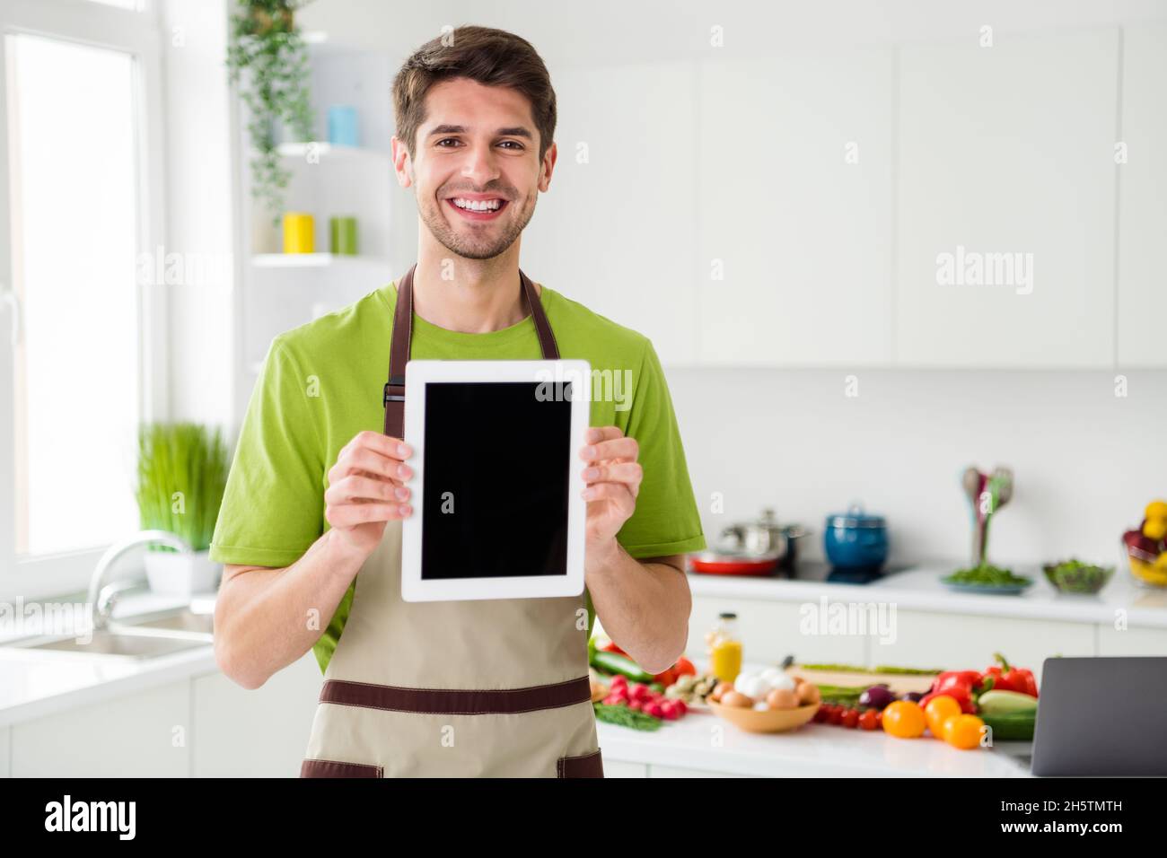 Ritratto di allegro ragazzo che cucina cena utilizzando il dispositivo che mostra schermo in casa cucina al chiuso Foto Stock
