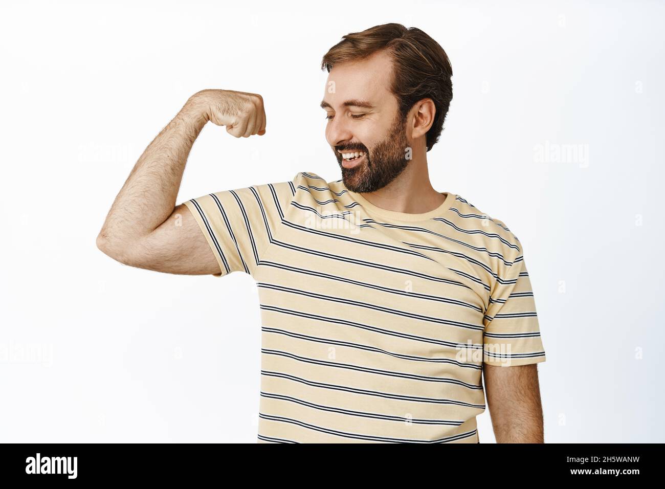 Ritratto di uomo sorridente compiaciuto guardando i muscoli del braccio, flettendo bicipiti con espressione soddisfatto del volto. Concetto di allenamento e di palestra Foto Stock