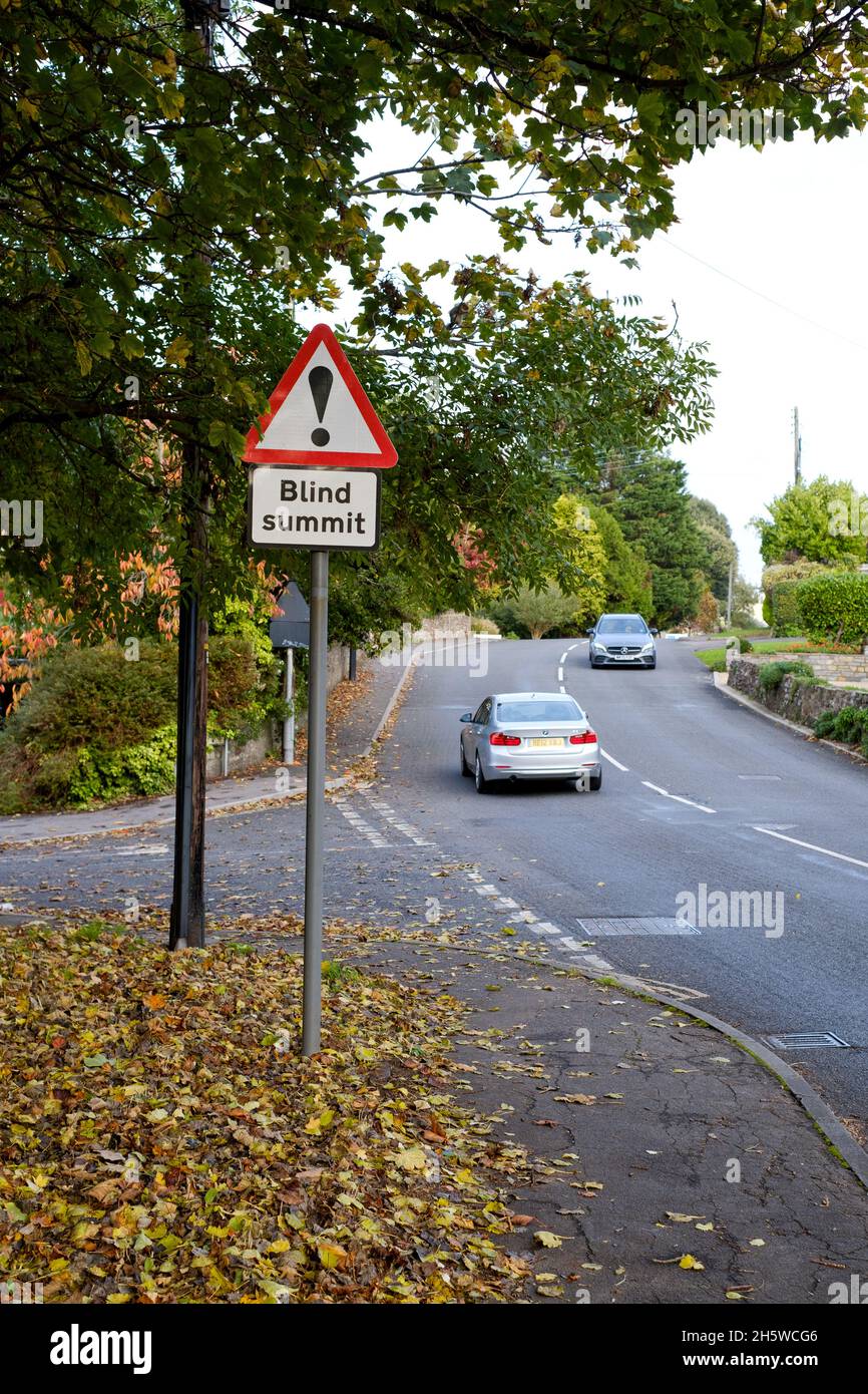 Un segnale di traffico britannico che avverte di un vertice cieco che precede è chiaramente visualizzato. Viene mostrato un veicolo che procede verso la sommità cieca su una strada residenziale Foto Stock