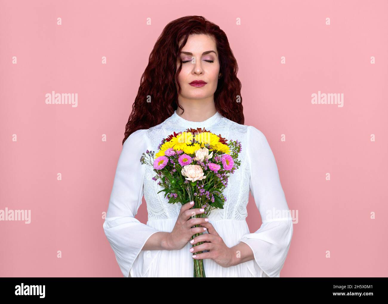 Donna con occhi chiusi tiene e odora il bouquet di fiori con sfondo rosa. Ritratto di bella donna in romantico abito bianco con fiori. Foto Stock
