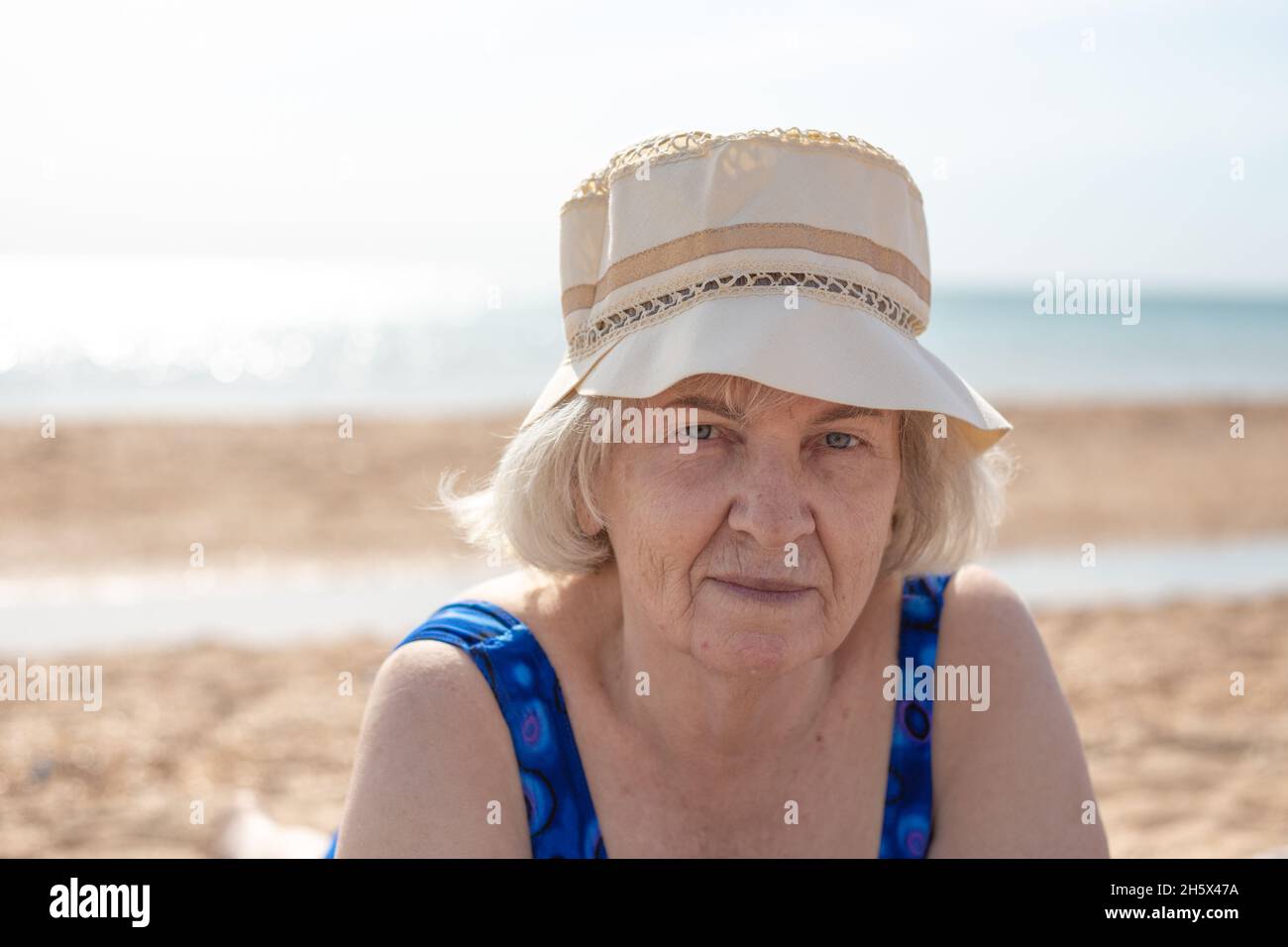 Una donna anziana in un cappello e un costume da bagno si trova sulla sabbia vicino al mare in una giornata estiva soleggiata. Riposo marittimo in pensione. Foto Stock