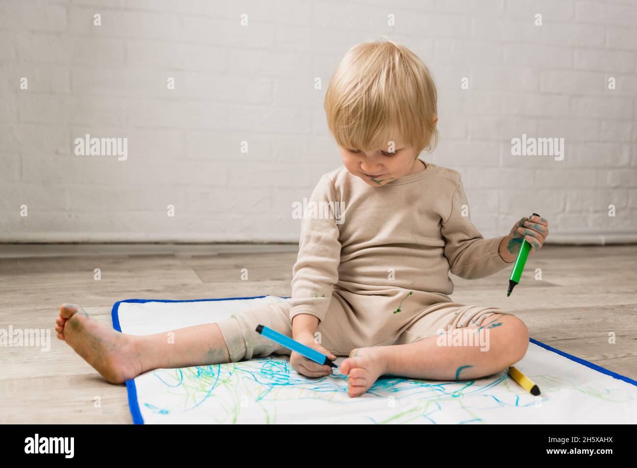 Una ragazza piccola allegra in pigiama è seduta sul pavimento e disegno con pennarello penne su uno speciale oilcloth. Viso e vestiti sono macchiati di dolore Foto Stock