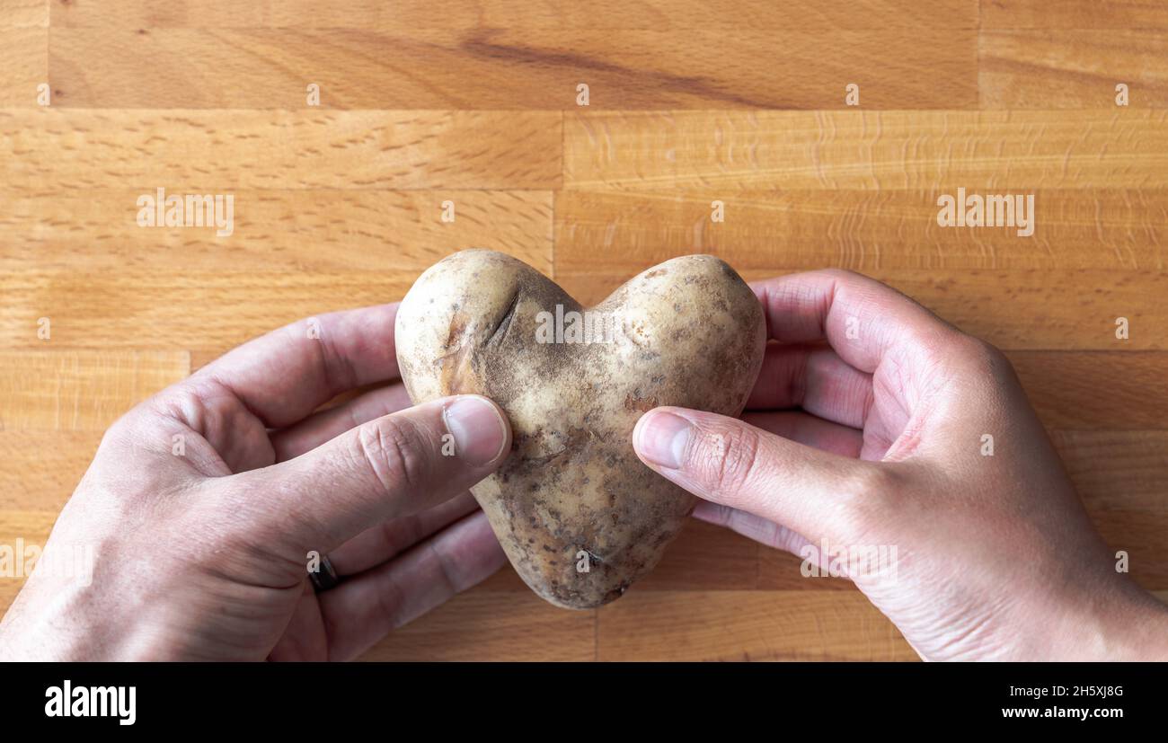 Un uomo e le mani di una donna condividono e tengono in mano una patata a forma di cuore su un tagliere di legno sfondo rendendo un romantico San Valentino amore backgr Foto Stock