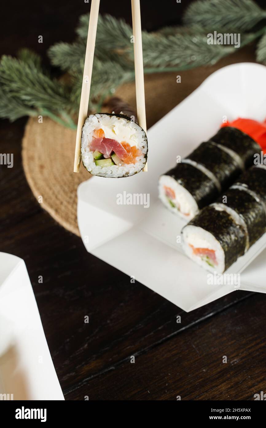 Tenere il rotolo di sushi utilizzando delle bacchette sul contenitore per la consegna degli alimenti. Decorazione di Natale. Consegna di cibo alla vigilia di Capodanno. Rotolare con salmone, tonno, cubum Foto Stock