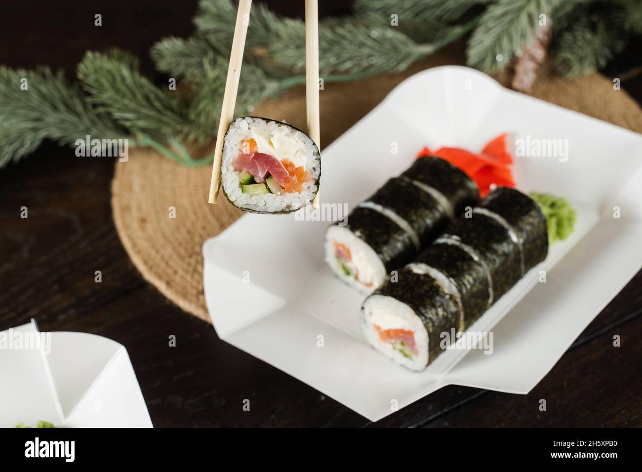 Tenere il rotolo di sushi utilizzando delle bacchette sul contenitore per la consegna degli alimenti. Decorazione di Natale. Consegna di cibo alla vigilia di Capodanno. Rotolare con salmone, tonno, cubum Foto Stock