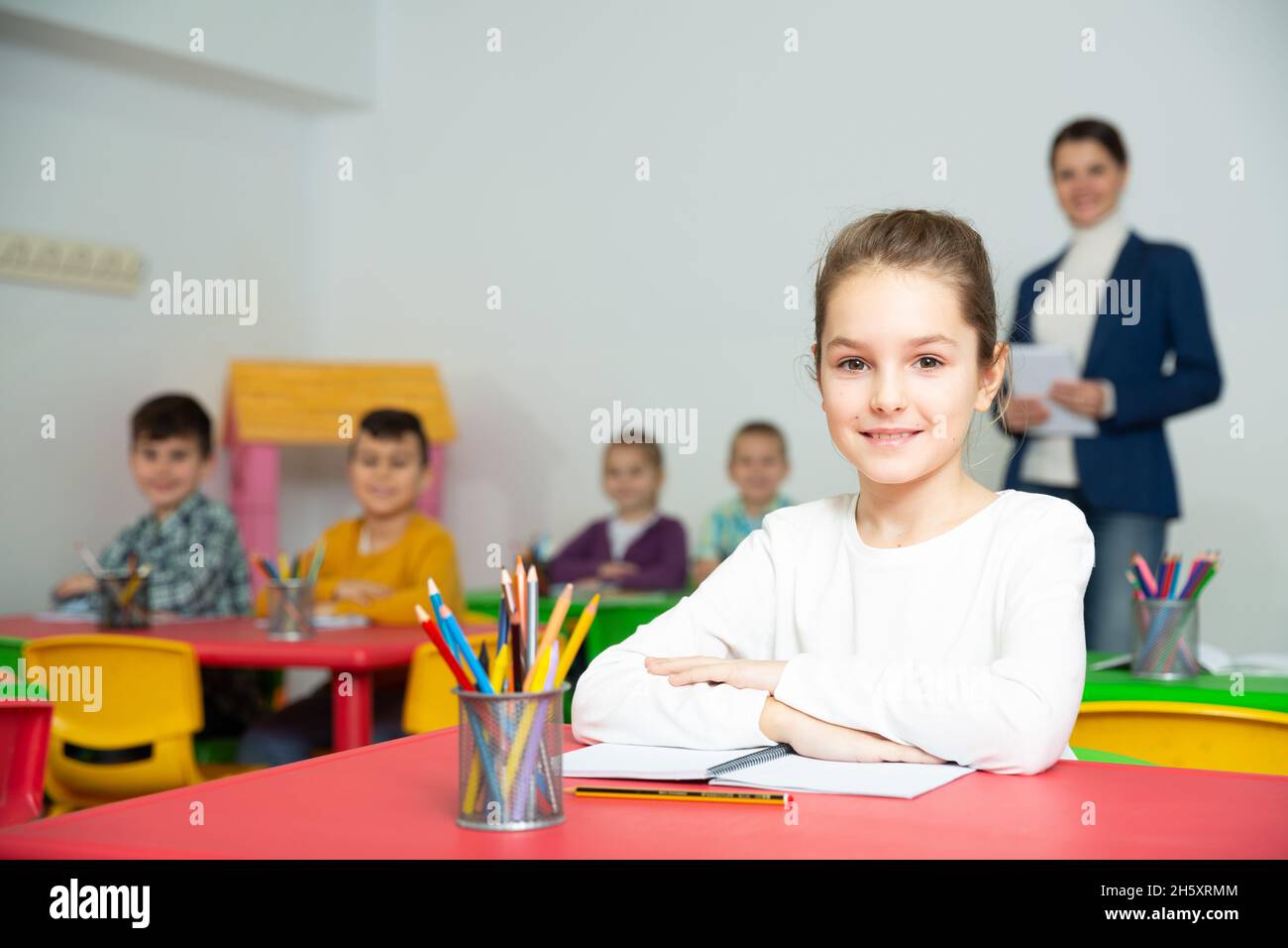 Carino ragazza di età che studia in classe su sfondo con compagni di classe e insegnante Foto Stock