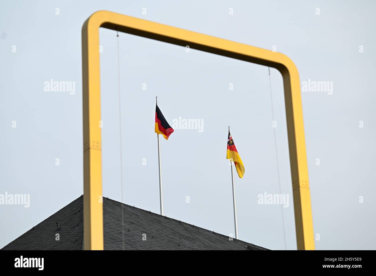 10 novembre 2021, Renania-Palatinato, Magonza: Un telaio in ottone vuoto dell'installazione 'tre colori' dell'artista berlinese Michael Sailstorfer si erge su un prato davanti alle bandiere del parlamento di Stato della Renania-Palatinato. L'opera d'arte di fronte al rinnovato parlamento di Stato dovrebbe essere completata la prossima settimana. I pannelli in tessuto alti sei metri e larghi tre metri in nero, rosso e oro erano già stati abbattuti alcune settimane fa come precauzione a causa di un avvertimento di tempesta. Da allora, i tre archi in metallo sono stati vuoti davanti al Deutschhaus. (A dpa 'bandiere per retur Foto Stock