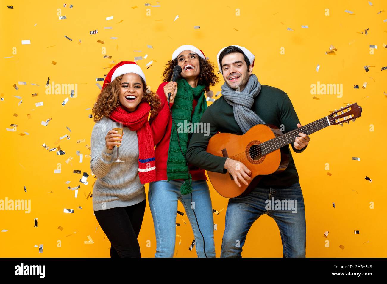 Gruppo di amici allegri e diversi che hanno partito cantando e festeggiando il Natale in giallo studio sfondo con confetti Foto Stock