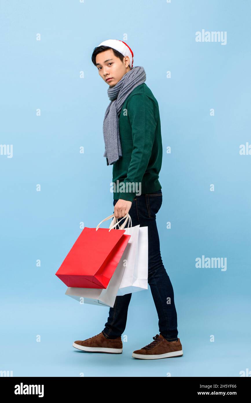 Handsosme uomo asiatico in tema di Natale attrie a piedi e tenere borse per lo shopping in uno studio di colore azzurro Foto Stock