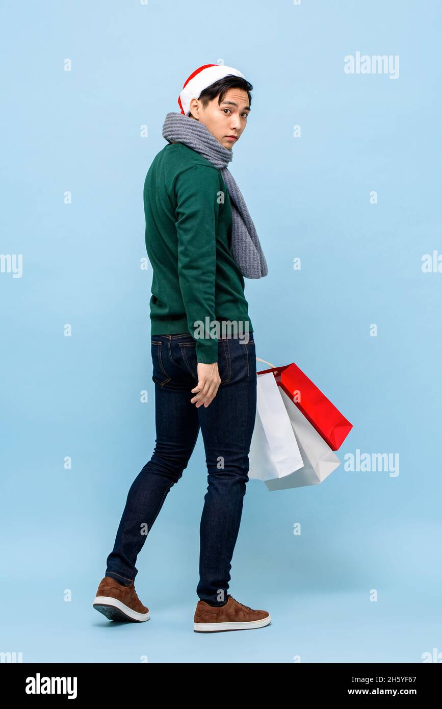 Handsosme uomo asiatico in tema di Natale attrie a piedi e tenere borse per lo shopping in uno studio di colore azzurro Foto Stock