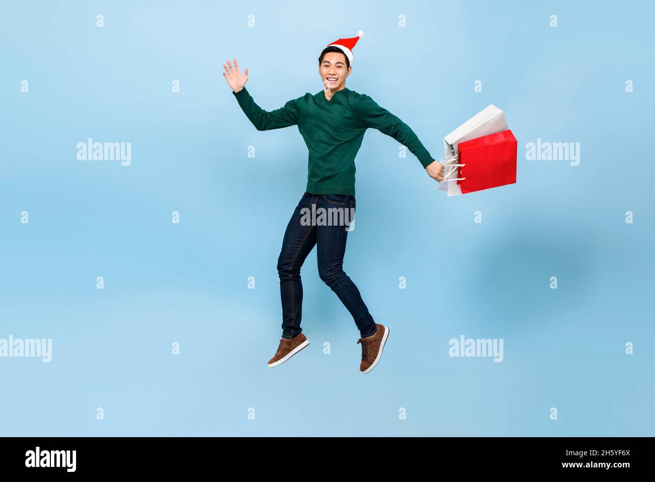 Ritratto a tutta lunghezza di felice giovane uomo asiatico bello indossare cappello di natale saltando mentre tenendo borse shopping isolato su sfondo azzurro chiaro Foto Stock