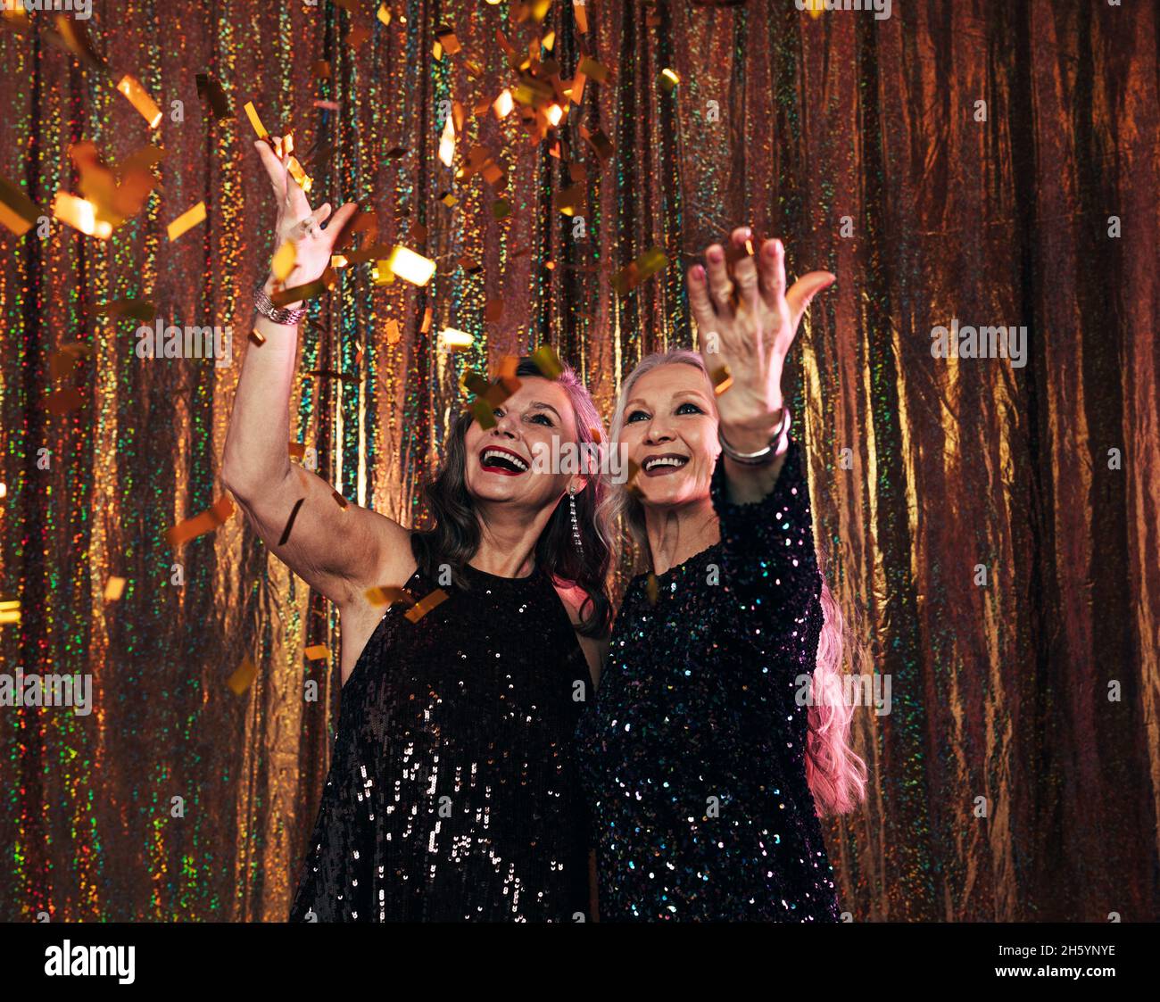 Due donne anziane sorridenti in abiti neri che lanciano confetti colorati mentre si ergono su uno sfondo dorato in studio Foto Stock