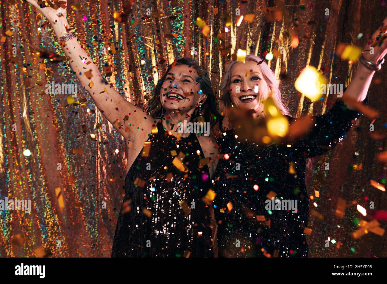 Due donne adulte felici in abiti neri che si divertono sotto confetti colorati in piedi su uno sfondo dorato Foto Stock