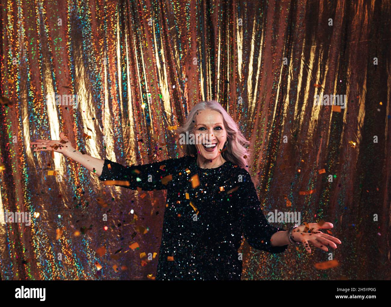 Donna anziana felice in abito nero in piedi sotto i confetti su uno sfondo dorato Foto Stock